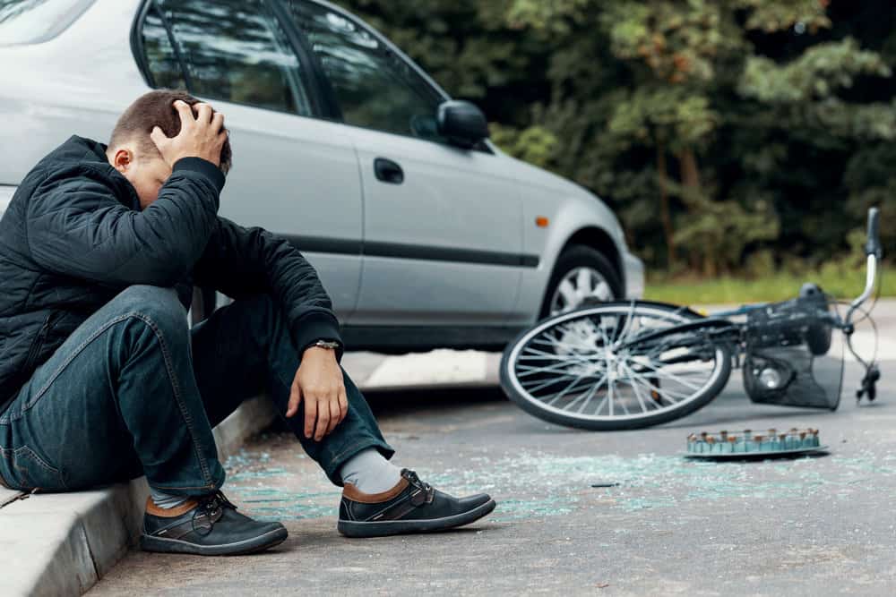 Worried biker holding his head and sitting on a pavement next to a car and bike crash